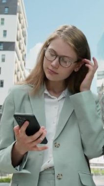 Gözlüklü genç iş kadınının dikey görüntüsü. Takım elbise giyiyor. Telefondaki haritaları kullanarak yerini buluyor ve dışarı bakıyor.