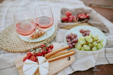 Piknik battaniyesinde gül şarabı, peynir, meyve ve atıştırmalık bardaklar. Güzelce düzenlenmiş peynir, üzüm, galeta ve domates parçaları yaz pikniği hazırlıyor.