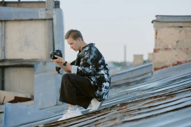 Çatıda otururken dijital kamerayı ayarlarken görülen şahıs, açık gökyüzü altında kentsel ortamda bir görüntü yakalamaya odaklanıyor