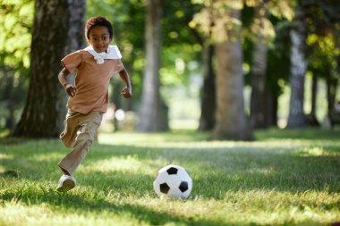 Parkta çimenlerde koşan Afrikalı Amerikalı çocuk, tek başına futbol oynuyor.