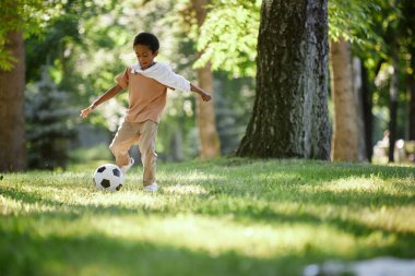 Küçük Afrikalı Amerikalı çocuk sıcak yaz gününde parkta futbol yeteneğini geliştiriyor.