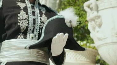 Süslü süvari üniformalı, beyaz eldivenli, beyaz tüylü şapkalı, dışarıda, tarihi binanın önünde dikilen, tanınmamış bir adamın yakından görüntüsü.