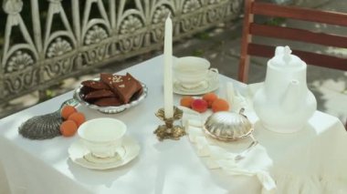 Beyaz porselen çaydanlık, çay fincanları, şekerli gümüş tepsi, taze kayısılar ve gün ışığında beyaz masa örtüsü üzerine mum.