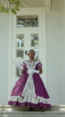 Dikey çekimde, klasik giysili ve eldivenli siyahi kadın elinde yelpaze ile tarihi binanın girişinde dikiliyor ve birini beklerken etrafına bakıyor.