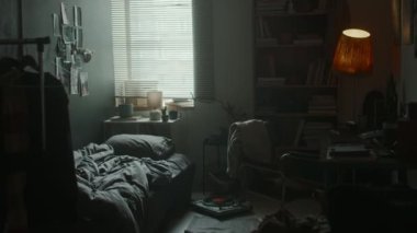 Loş ışıklı odanın içinde panjurları, duvar posterleri, koyu gri çarşaflı yapılmamış yatak, deri koltuklu masanın yanında lamba, yerde plak çalar ve uzun kitaplık var.