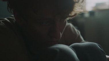 Üzgün genç bir adamın boş gözlerle bakışlarını yakın plan çekimleri, sonra odasında yerde otururken dizlerinin üzerinde dinlenmesi.