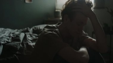 Uykucu genç adam dizlerinin üstüne çöker, elleri saçlarının arasında koşar, yanaklarını ovuşturur ve odasında dağınık bir şekilde yatar.