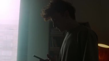 Tereddütlü genç adam telefona bakıyor, sonra yapılmamış yatağa oturuyor ve yansıması için aletleri bir kenara atıyor.