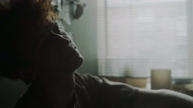 Depresyonda olan genç adamın yakın çekim görüntüsü, duvara yaslanmış kafası ve pencereye yaslanmış sigara içişi.