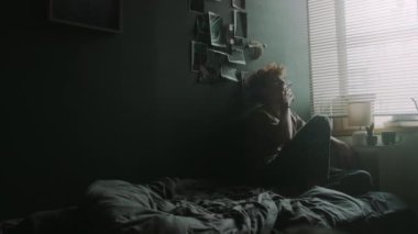 İçine kapanık genç adam pencerenin yanında dağınık bir yatakta oturuyor. Panjurları var ve siyah duvara yaslanmış sigara içiyor.