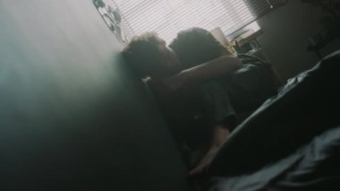 Hollanda bakış açısına göre, gergin genç adam, kollarını kavuşturup dizlerini bükerek yatakta otururken gri duvara vuruyor.