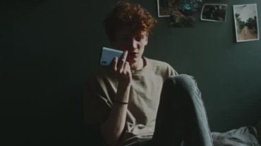 Endişeli genç adam telefonda konuşurken hoparlör modunda yatakta oturup duvara yaslanıyor.