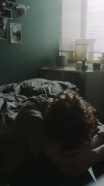 Dikey çekimde, genç bir adam başını dizlerinin üstüne koyup boynuna dokunurken dağınık bir yatağın yanında oturuyor.