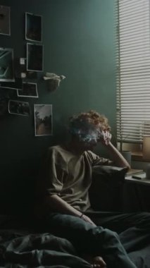 Yatak odasında panjurlu yatağın yanında sigara içerken, gündelik giysiler içinde perişan bir genç adamın dikey çekimi.