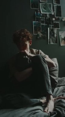 Evinde, karanlık duvarların karşısında oturmuş yastığa sarılan endişeli genç adamın dikey görüntüsü.