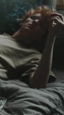 Yatakta uzanmış sigara içen, kültablası dolusu izmariti olan depresif bir gencin dikey görüntüsü.