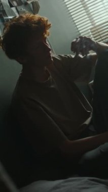 Mutsuz genç bir adamın yatak odasında karanlık duvarın karşısında sigara içişini dikey olarak görmek.