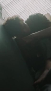 Yatakta pencerenin yanında panjurlarla oturan hareketli genç adamın dikey görüntüsü.