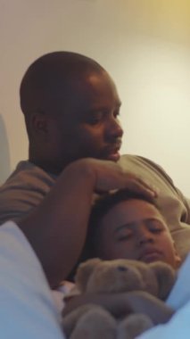 Afro-Amerikan babasının göğsünde yatıp sesli okuduğu ve oğullarının kafasını okşadığı uykulu küçük çocuğun dikey görüntüsü.