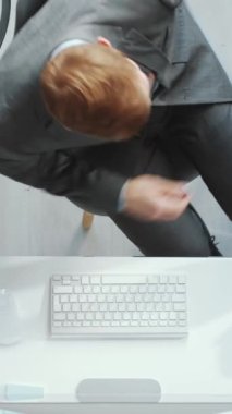 Erkek çalışanın iş yerine doğru yürüdüğü, sandalyeye oturduğu, telefonu masaya koyduğu, not defterini çantasından çıkardığı ve bilgisayarla çalışmaya başladığı dikey kare.