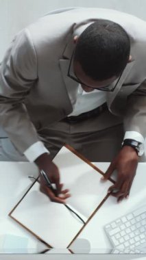 Ofis masasında çalışırken bilgisayar kullanan ve not defterinde yazan takım elbiseli Afro-Amerikan işadamının dikey aşağı bakışı