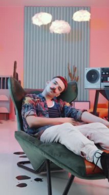 Dikey çekimde, tuhaf bıyıklı adam, klasik getto silahının yanındaki koltukta dinleniyor, kameraya bakıyor ve parlak renkli ışık saçan moda stüdyoda moda el hareketleri yapıyor.