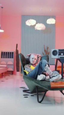 Vibey kısaltmalı, modaya uygun kıyafetleri olan, retro müzik kutusunun yanındaki koltukta dinlenen, müzik dinleyen ve pop sanat dekoru olan bir odada kameraya poz veren bir kızın dikey görüntüsü.
