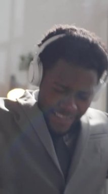 Formalite icabı giyinen genç siyahi işadamının kablosuz kulaklık taktığı, favori müziği dinlediği, güneşli ofiste otururken heyecanla şarkı söylediği ve dans ettiği dikey bir fotoğraf.