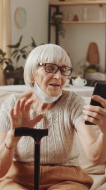 Çene ve baston altında koruyucu maskeli yaşlı bir bayanın kameraya el sallarken akıllı telefonu tutarken ve evde video görüşmesi yaparken dikey görüntüsü.