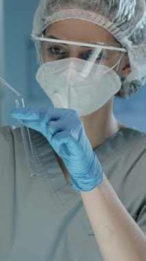 Dikey olarak, koruyucu giysiler içindeki genç bayan doktorun deney tüpüne pipetle sıvı döküp laboratuvarda incelemesi.