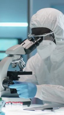 Koruyucu giysiler içinde Afrikalı Amerikalı erkek biyoteknoloji uzmanının laboratuvarda mikroskopla örneğe baktığı dikey görüş.