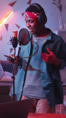 Genç Afrikalı Amerikalı bir adamın kulaklıkla telefon ekranından şarkı okuyup mikrofona vururken evde yeni şarkılar kaydettiği dikey bir çekim.