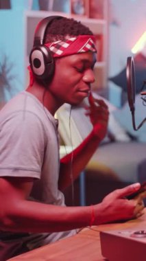 Dikey çekimde siyah rapçi kulaklık takıyor telefonda şarkı söylüyor ve mikrofona şarkı söylüyor. Ev stüdyomuzda renkli neon ışıklandırmayla yeni bir şarkı kaydediyor.