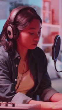 Ev kayıt stüdyosundaki mikrofon ve ses panosuyla kameraya gülümseyen ve poz veren Asyalı genç bayan müzisyenin dikey portresi.