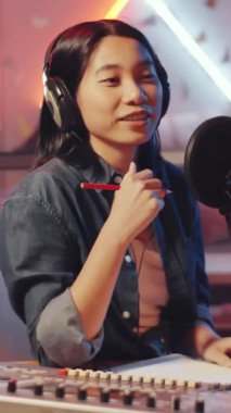 Kulaklıkla Asyalı kadın podcaster 'ın mikrofona konuşurken renkli ışıklandırmalı ses stüdyosundaki konukla yaptığı röportajın dikey görüntüsü.