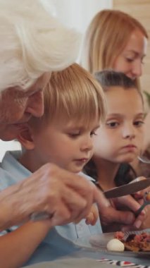 Büyükannenin çatal bıçakla yemek keserken aile yemeği sırasında küçük çocuğa yardım ettiği dikey görünüm, arka plandaki kız ve kadın