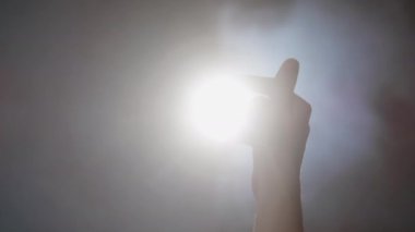 Nazikçe hareket eden kadın elinin yakın plan görüntüsü, parmaklar arasındaki boşluklardan parlayan parlak ışık kaynağı, sağdan sola üfleyen beyaz duman, umut ve arzu kavramı.