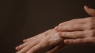 Kimliği belirsiz bir kadının avucuna krem sürerken ve koyu kahverengi arka planda el derisine sürerken yakın plan fotoğrafı.