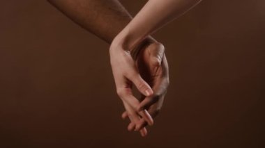 Çok ırklı çiftin birbirine kenetlenmiş parmaklarıyla el ele tutuşup kahverengi arka planda birlik gösterdikleri yakın plan görüntüsü