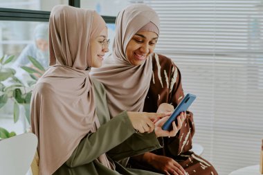 İki Müslüman kadın akıllı telefona bakarken gülümsüyor, modern içteki bitkilerle çevrili, başörtüsü ve şık kıyafetler giyip, birlikte anlarını paylaşıyor ve kaynaşıyorlar.