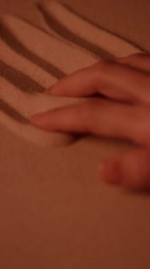 Kadın elinin dikey yakın çekimi. Sıcak kumda dört parmağıyla çizgiler çiziyor.