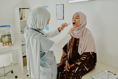 Tıbbi uzman, iyi donanımlı bir klinikte hastaların boğazını muayene ediyor. Hasta sakince tesettüre oturuyor, hemşire özel tıbbi ekipman kullanıyor, ön planda pano var.