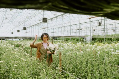 Serada çiçek buketi tutan, şapka ve gözlük takan yemyeşil çiçeklerle çevrili biri. Gülümseyen ve dingin ortamın tadını çıkaran