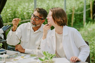 Two scientists analyzing plant samples outdoors, holding test tube and discussing results, surrounded by laboratory equipment and greenery under a shade structure clipart