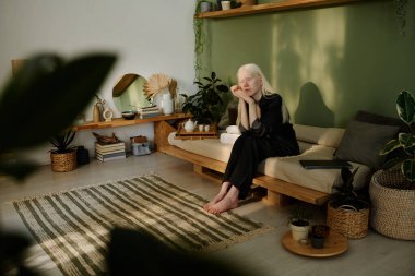 Geniş açılı, albinizmli genç beyaz kız oturma odasında, modern Wabi Sabi iç mekanıyla rahatça oturuyor.