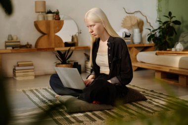 Albinizmli genç Z jenerasyonlu kadın Wabi Sabi oturma odasında yerde oturuyor ve modern dizüstü bilgisayarında daktilo yazıyor.