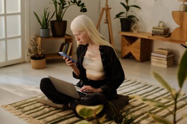 Beyaz saçlı, uzun beyaz saçlı genç bir kadın Wabi-Sabi oturma odasında oturuyor. Sabah laptopuyla çalışırken akıllı telefondan mesaj gönderiyor.