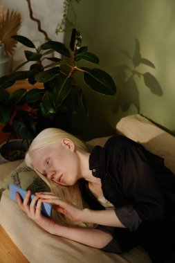 Albinizmli beyaz bir kızın kanepede dinlenip akıllı telefondan video izlediği dikey yüksek açı görüntüsü.