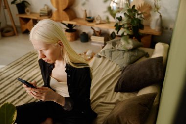 Samimi oturma odasında oturan albinizmli genç bir kadının yüksek açılı görüntüsü akıllı telefondan sosyal medyayı çekiyor, uzayı kopyalıyor.