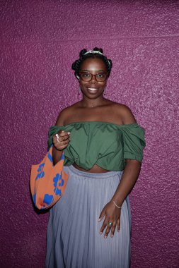 Sevimli Afro-Amerikan kadın şık giyinmiş geniş gülümsemeli ve arkasında mor bir duvar olan portre için poz veriyor.